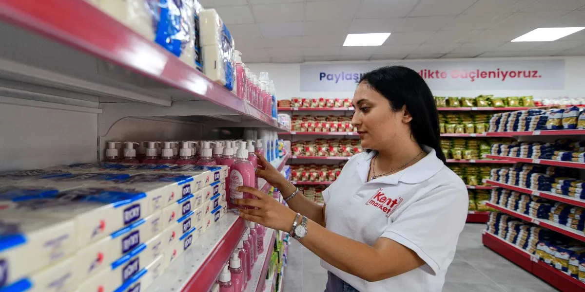 Kent Market logolu beyaz tişört giyen bir kadın, market rafındaki sıvı sabunları düzenliyor. Arka planda raflarda gıda ürünlerinin dizili olduğu marketin duvarında 