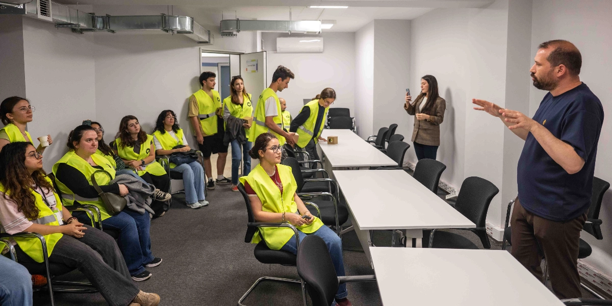 Kapalı bir toplantı odasında, üzerinde neon sarı yelekler bulunan bir grup genç, ayakta duran ve konuşma yapan sakallı bir adamı dikkatle dinliyor. Gençlerin çoğu sandalyelerde otururken, bazıları ayakta duruyor ve odanın kenarında bir kadın cep telefonuyla fotoğraf çekiyor. Ortada uzun beyaz masaların bulunduğu odada, katılımcılar ellerinde küçük karton kutularla bir etkinlik veya eğitim sürecinde görünüyor.
