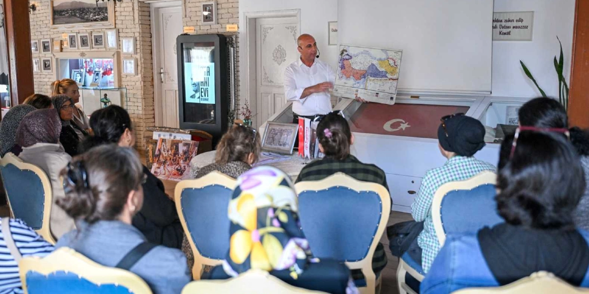 Tarihi bir mekanda, beyaz gömlekli bir erkek, önünde oturan kadın ve çocuklardan oluşan bir gruba elindeki Türkiye haritasını göstererek sunum yapıyor. Dinleyiciler mavi döşemeli sandalyelerde oturuyor, arkadaki masanın üzerinde ise Türk bayrağı ve çeşitli tarihi görseller yer alıyor. Duvarlarda çerçevelenmiş fotoğraflar ve üzerinde yazı bulunan bir levha bulunuyor.