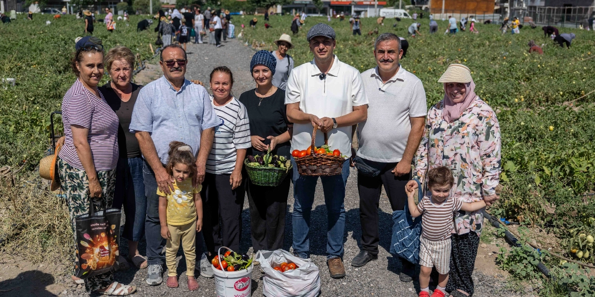 Doğanlar Kent Bostanı'nda hasat yapan bir grup insan, topladıkları sebzelerle birlikte tarlanın içindeki toprak yolda yan yana duruyor. Ön planda, ellerinde hasat ettikleri domates ve patlıcan dolu sepetler, plastik bir kova ve poşet bulunan yetişkinler ile yanlarında küçük çocuklar yer alıyor. Arka planda ise geniş bir alana yayılmış sebze tarlası ve hasada devam eden diğer insanlar görülüyor.