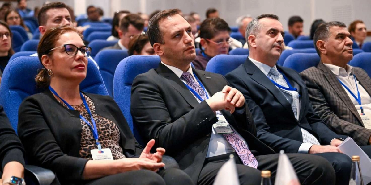 Konferans salonunda mavi koltuklarda oturan, boyunlarında etkinlik kartı asılı olan ve dikkatle bir konuşmayı dinleyen katılımcılar. Ön sırada soldan sağa doğru gözlüklü bir kadın, yanında desenli kravatlı bir erkek ve takım elbiseli diğer katılımcılar yer alıyor.