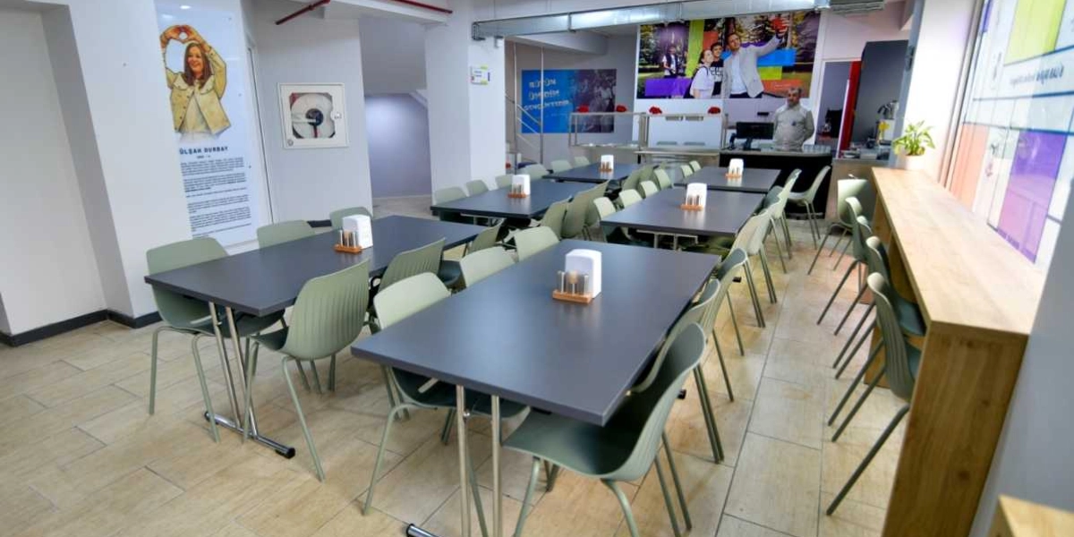 Modern bir yemekhane veya kafe iç mekanı; gri masalar, etrafında açık yeşil plastik sandalyeler, sağ tarafta ahşap bir bar tezgahı ve duvarlarda belediye faaliyetlerini gösteren görseller ile bilgilendirme panoları bulunuyor.