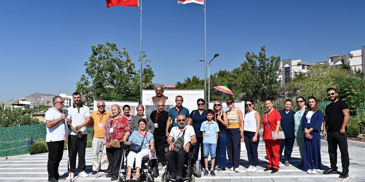 Açık havada, arkalarında Türk bayrağı ve Atatürk büstü bulunan, farklı yaş ve fiziksel özelliklere sahip yaklaşık yirmi kişilik bir grup, güneşli bir günde yan yana durarak poz veriyor. Grubun ön sırasında tekerlekli sandalye kullanan iki kişi yer alırken, diğer katılımcılar ayakta duruyor. Aralarında beyaz baston kullanan bir kişinin de bulunduğu grup, günlük kıyafetler içinde, park benzeri bir alanda, binaların ve ağaçların önünde bir arada bulunuyor.