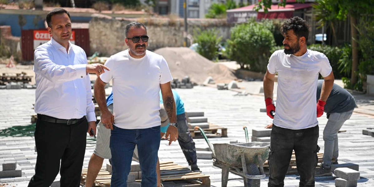 Beyaz gömlekli ve siyah pantolonlu bir yetkili, inşaat halindeki bir alanda yanındaki kişiye eliyle yön gösteriyor. Yanında beyaz tişörtlü ve güneş gözlüklü bir adam ile sağ tarafta kırmızı eldivenli, beyaz tişörtlü bir işçi bulunuyor. Arka planda ise zemin döşeme çalışmaları devam eden bir meydan ve çalışan diğer işçiler yer alıyor.