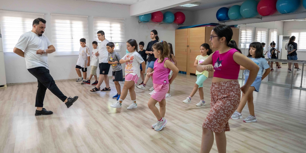 Beyaz tişörtlü bir eğitmen eşliğinde dans veya hareket eğitimi alan bir grup çocuk, geniş ve aynalı bir spor salonunda aynı adımları atarak pratik yapıyor.