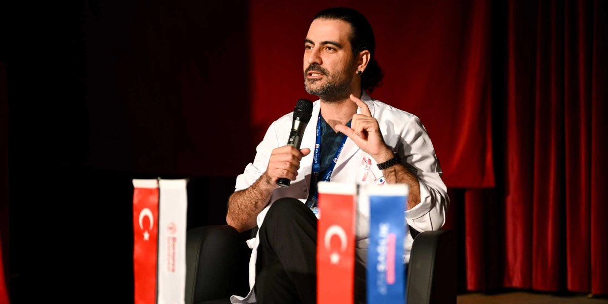 Kırmızı perde önünde siyah bir koltukta oturan, beyaz önlüklü bir erkek doktor, elinde mikrofonla konuşma yapıyor. Doktorun önlüğünün üzerinde meme kanseri farkındalığını simgeleyen pembe kurdele takılı. Ön planda, üzerinde Türk bayrağı ve kurum logolarının bulunduğu dikey pankartlar yer alıyor.