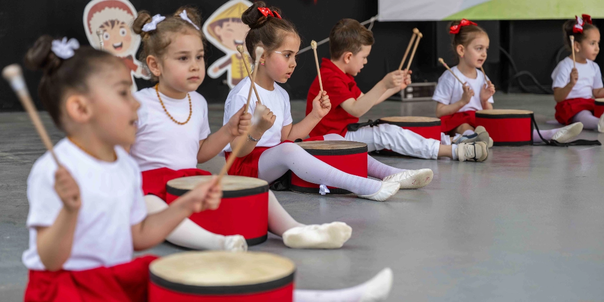 Beyaz tişört ve kırmızı etek veya pantolon giymiş bir grup çocuk, sahnede önlerinde duran kırmızı davullara bagetlerle vurarak ritim tutuyor. Çocukların bir kısmı saçlarını topuz yapmış, bazıları ise kırmızı kurdeleli tokalar takmış durumda. Arka planda çocuk figürlerinin olduğu dekoratif panolar yer alıyor.