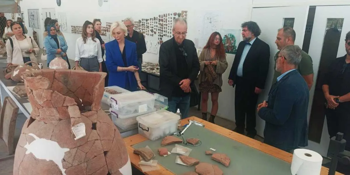 Aydınlık bir müze veya laboratuvar ortamında, masaların üzerinde sergilenen arkeolojik çanak çömlek parçalarını ve büyük bir pişmiş toprak küpü inceleyen bir grup insan. Ön planda, parçaları birleştirilmiş büyük bir antik küp ve masanın üzerinde düzenli şekilde dizilmiş seramik parçaları yer alıyor. Aralarında resmi kıyafetli yetkililerin ve araştırmacıların bulunduğu grup, dikkatle eserleri incelerken birbirleriyle iletişim kuruyor. Arka plandaki beyaz duvarlarda ise arkeolojik bulguları gösteren çizimler ve belgeler asılı duruyor.
