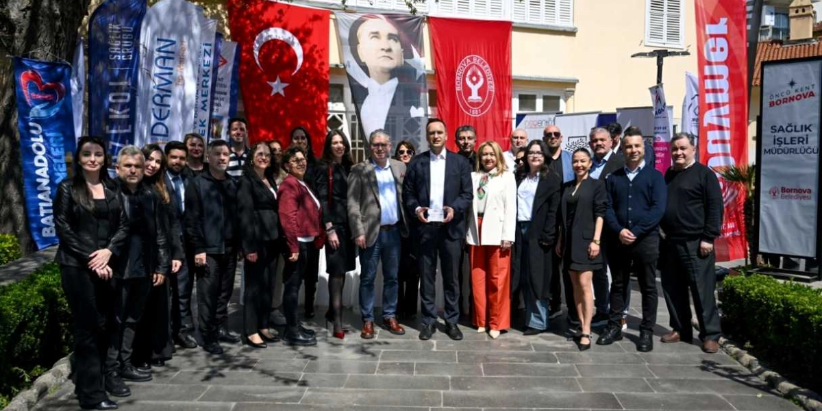 Bornova Belediyesi önünde, arkalarında Türk bayrağı, Atatürk posteri ve çeşitli kurumların flamalarının yer aldığı bir binanın önünde yan yana duran, resmi ve günlük kıyafetli yaklaşık yirmi kişilik bir grup. Grubun merkezinde yer alan bir erkek, elinde küçük bir plaket tutuyor. Sağ tarafta üzerinde 