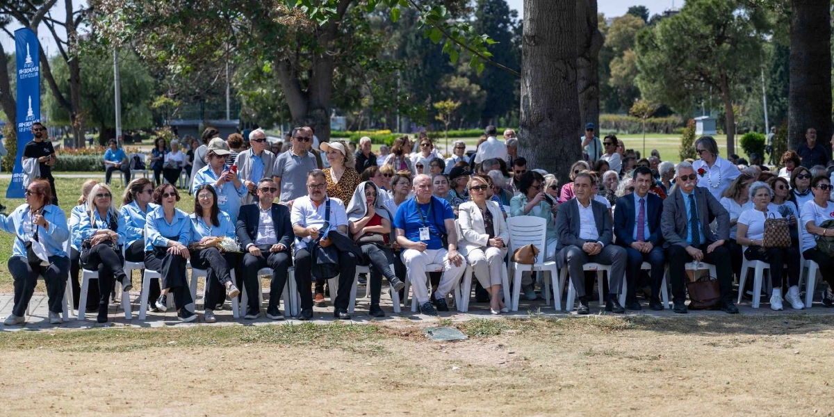 Açık havada, ağaçların gölgesindeki bir alanda beyaz plastik sandalyelerde yan yana oturan, çoğunluğu orta ve ileri yaş grubundaki katılımcılardan oluşan bir topluluk. Katılımcıların bir kısmı açık mavi gömlekler giymiş, bir kısmı ise resmi kıyafetlerle etkinliği takip ediyor. Arka planda İzmir Büyükşehir Belediyesi logosunun yer aldığı mavi bir flama bulunuyor.