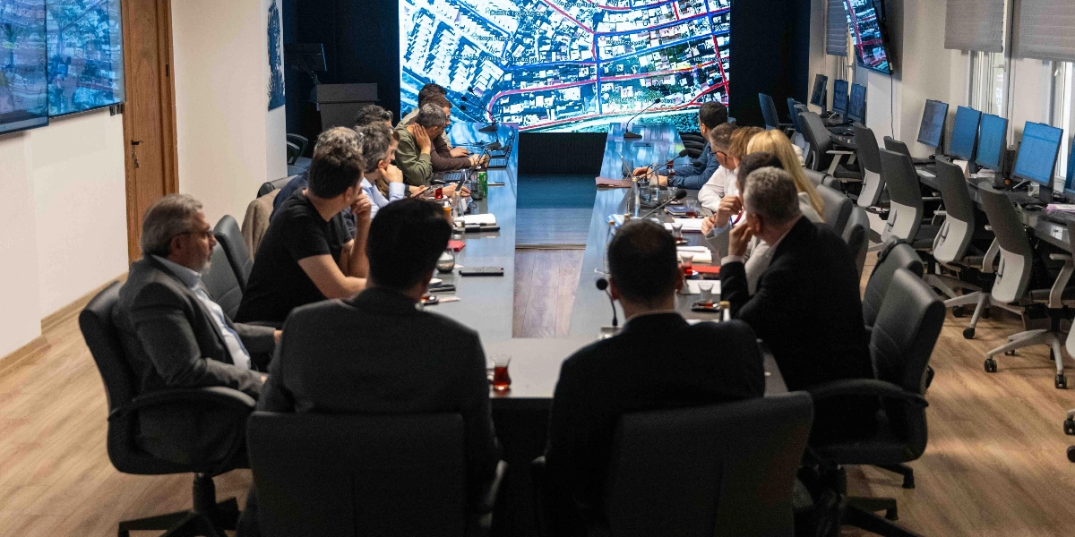 Modern bir toplantı odasında, uzun bir masa etrafında toplanmış, dijital harita ve veri görselleştirmelerinin yansıtıldığı büyük bir ekranı inceleyen bir grup profesyonel. Odanın yan tarafında bilgisayar istasyonları bulunurken, masada oturan katılımcılar ekranı takip ederek toplantılarını sürdürüyor.