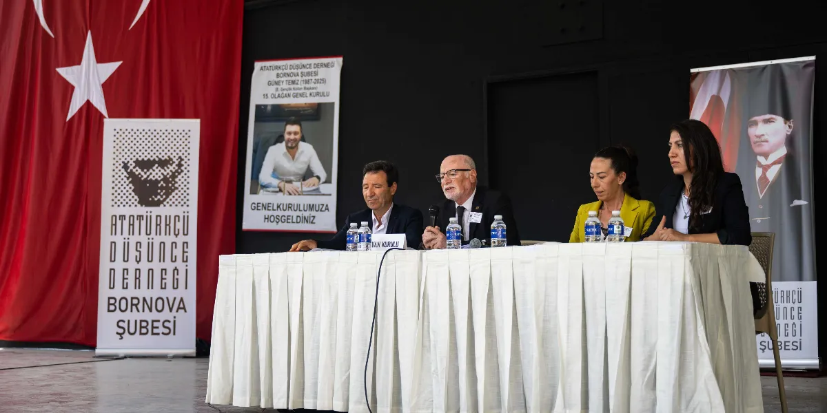 Atatürkçü Düşünce Derneği Bornova Şubesi 15. Olağan Genel Kurulu'nda, beyaz örtülü bir masada oturan dört kişilik divan heyeti görülüyor. Masanın ortasında mikrofonla konuşan gözlüklü bir beyefendi, yanında diğer kurul üyeleri yer alıyor. Arka planda solda büyük bir Türk bayrağı, ortada hayatını kaybeden Gençlik Kolları Başkanı Güney Temiz'in anısına hazırlanan 