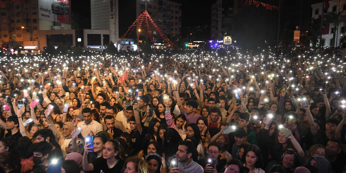 Gece vakti Bornova'da düzenlenen Cumhuriyet Bayramı kutlamasında, kalabalık bir insan grubu ellerindeki telefonların ışıklarını havaya kaldırarak alanı aydınlatıyor. Arka planda şehir ışıkları ve binalar görünürken, ön planda coşkulu bir şekilde kutlamaya katılan çok sayıda genç ve yetişkin yer alıyor.