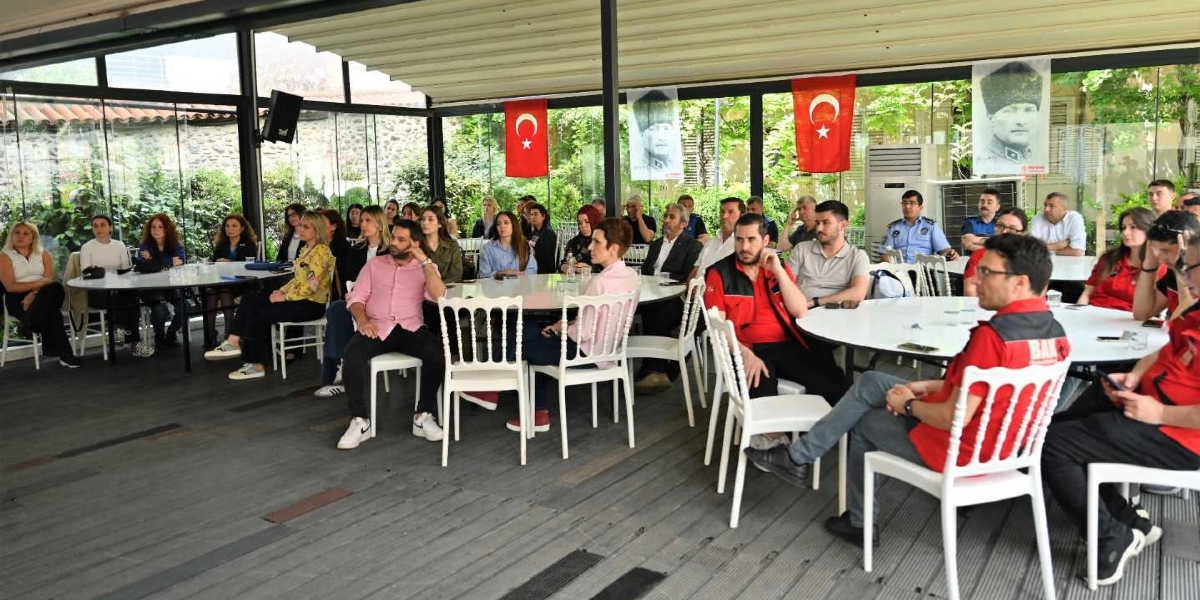 Açık hava bir mekanda, yuvarlak masalar etrafında toplanmış, dikkatle dinleyen bir grup katılımcı yer alıyor. Aralarında kırmızı yelekli arama kurtarma görevlilerinin de bulunduğu katılımcılar, sunumu takip ediyor. Mekanın arka duvarında asılı Türk bayrakları ve Mustafa Kemal Atatürk'ün portreleri bulunuyor. Ortam, resmi bir bilgilendirme veya eğitim toplantısını yansıtıyor.