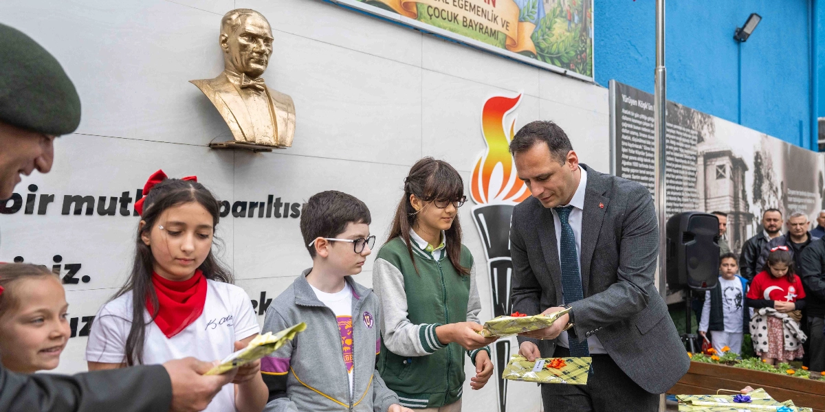 Bornova Belediye Başkanı, 23 Nisan Ulusal Egemenlik ve Çocuk Bayramı etkinlikleri kapsamında çocuklara hediye paketleri veriyor. Arka planda Mustafa Kemal Atatürk'ün büstü ve üzerinde 