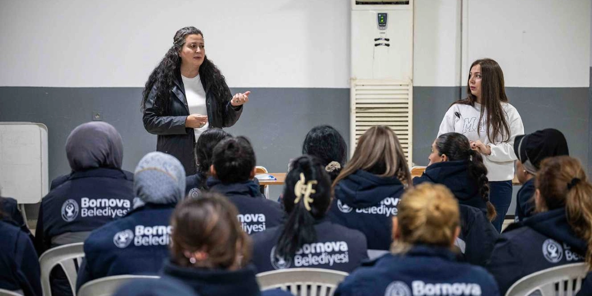 Kapalı bir mekanda, üzerinde Bornova Belediyesi yazılı lacivert ceketler giymiş kadınlardan oluşan bir gruba hitap eden bir kadın konuşmacı. Konuşmacı ayakta durarak elleriyle jestler yaparken, dinleyiciler sandalyelerde oturarak onu dinliyor. Sağ tarafta ise beyaz sweatshirt giymiş başka bir kadın daha bulunuyor.