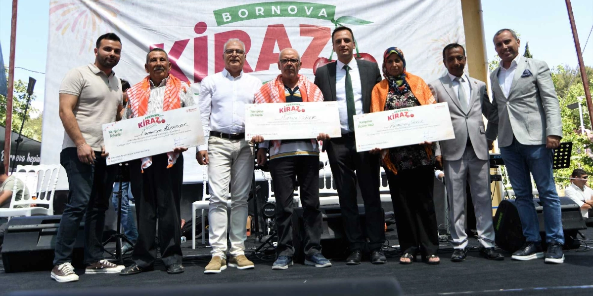 Bornova Kiraz Festivali kapsamında düzenlenen yarışmada dereceye giren üç kişi ve protokol üyeleri, üzerinde festival logosunun bulunduğu büyük bir pankartın önünde ödül çekleriyle poz veriyor. Soldan sağa doğru; genç bir erkek, omuzlarında kırmızı beyaz desenli şal olan yaşlı bir adam, beyaz gömlekli bir beyefendi, yine omuzlarında aynı şaldan olan başka bir yaşlı adam, koyu renk takım elbiseli ve yeşil kravatlı bir yetkili, başında eşarbı olan bir kadın, gri takım elbiseli bir beyefendi ve yine gri takım elbiseli bir başka beyefendi yan yana duruyor. Yarışmacılar ellerinde 