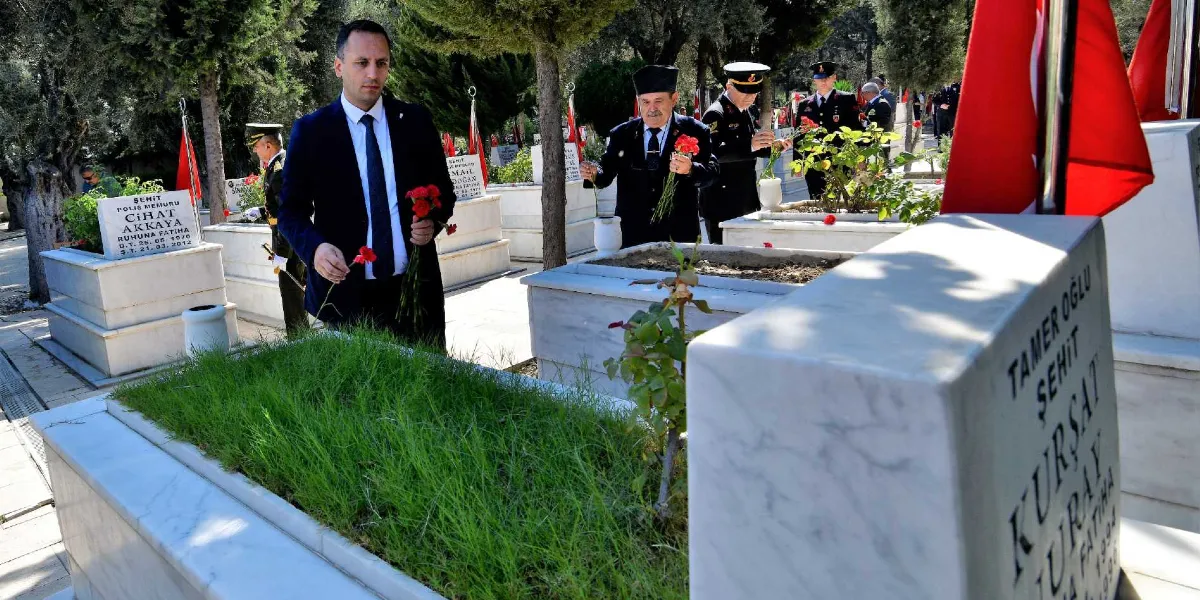 30 Ağustos Zafer Bayramı kapsamında bir şehitlikte düzenlenen anma töreninde, takım elbiseli bir yetkili ve askeri üniformalı kişiler, mermer mezar taşlarının bulunduğu alanda ellerinde kırmızı karanfillerle şehit kabirlerini ziyaret ediyor. Ön planda üzerinde 