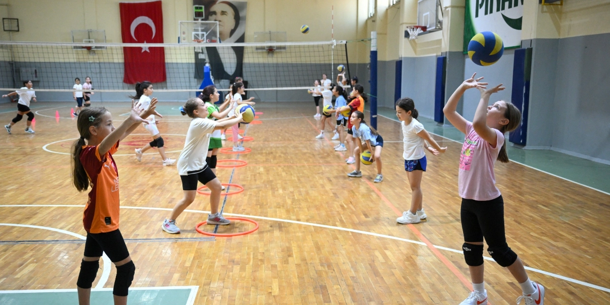 Kapalı bir spor salonunda voleybol antrenmanı yapan kız çocukları, yerdeki kırmızı çemberlerin etrafında sıralanmış şekilde voleybol toplarıyla pas çalışması yapıyor. Arka planda duvarda asılı Türk bayrağı ve Atatürk posteri görülüyor.