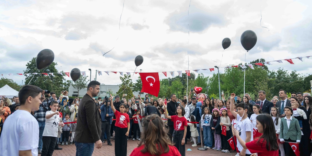 Açık havada düzenlenen bir etkinlikte, ellerinde Türk bayrakları ve iplerine bağlı siyah balonlar tutan çocuklar ve onları izleyen kalabalık bir grup yer alıyor. Çocukların bir kısmı üzerinde Atatürk baskılı kırmızı tişörtler giyerken, bir kısmı beyaz tişörtler tercih etmiş. Arka planda Türk bayrakları ve süslemelerle donatılmış bir meydan, ön planda ise çocuklara eşlik eden yetişkinler görülüyor. Gökyüzüne doğru bırakılan balonlar ve çocukların neşeli tavırları, kutlama havasını yansıtıyor.