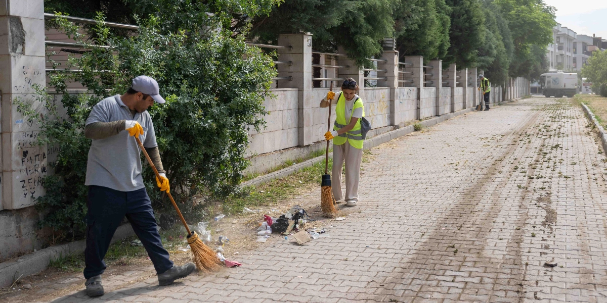 Bornova'da bir sokakta temizlik yapan belediye görevlileri, ellerindeki süpürgelerle kaldırımdaki çöpleri bir araya getiriyor. Sarı reflektörlü yelek giymiş görevliler, parke taşlı yolda çalışırken arka planda bir temizlik aracı ve ağaçlıklı bir duvar hattı görülüyor.