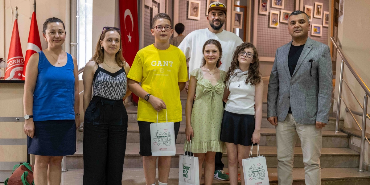 İç mekan merdivenlerinde yan yana duran, aralarında çocukların ve yetişkinlerin bulunduğu yedi kişilik bir grup. Gruptaki üç çocuk ellerinde üzerinde Bornova Belediyesi logosu ve çizimleri bulunan beyaz hediye çantaları tutuyor. Arka planda Türk bayrağı ve duvarda sergilenen fotoğraflar yer alıyor. Çocuklar ve yetişkinler gülümseyerek kameraya bakıyor.