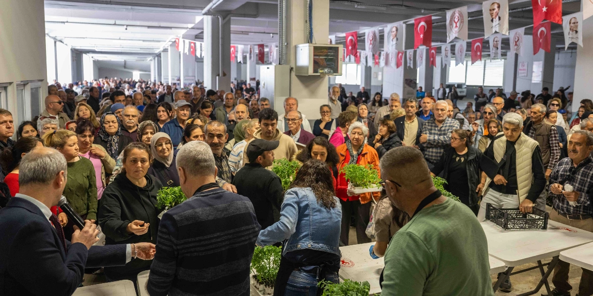 Kapalı bir alanda düzenlenen etkinlikte, Bornova Belediyesi yetkilileri tarafından vatandaşlara ücretsiz fide dağıtılıyor. Kalabalık bir insan grubu, masaların etrafında toplanmış, ellerinde küçük saksılarda yeşil fidelerle bekliyor. Ön planda bir yetkili mikrofonla konuşurken, diğer görevliler masaların üzerindeki fide kasalarını dağıtıyor. Tavandan sarkan Türk bayrakları ve Atatürk posterleriyle süslenmiş geniş bir depo veya pazar yeri atmosferi hakim.