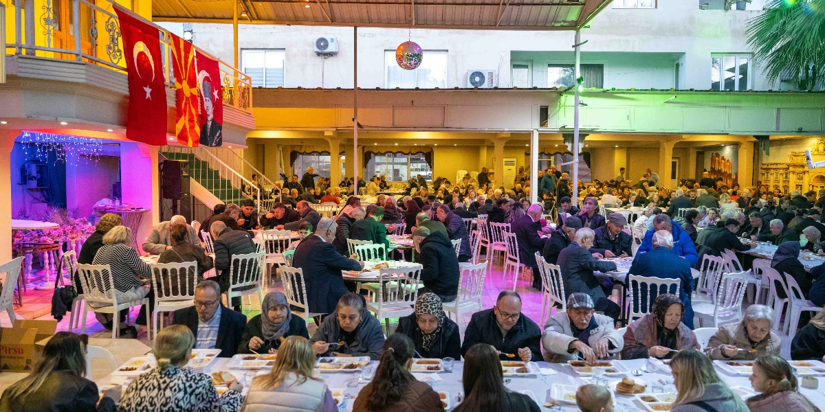 İzmir Makedonya Göçmenleri Kültürünü Yaşatma ve Dayanışma Derneği'nin düzenlediği iftar yemeğinde, Bornova Belediye Başkanı Ömer Eşki'nin de aralarında bulunduğu çok sayıda vatandaş, açık hava alanında kurulan masalarda birlikte yemek yiyor. Arka planda asılı Türk bayrağı ve Makedonya bayrağı ile Mustafa Kemal Atatürk'ün portresi dikkat çekerken, katılımcılar beyaz örtülü masalarda oturarak iftar yapıyor.