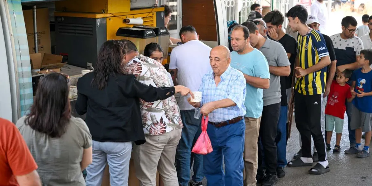 Bornova Belediyesi'ne ait bir ikram aracının önünde, yaşlı bir adamın elinde plastik bir kapla yemek aldığı ve arkasında farklı yaş gruplarından insanların sıra beklediği bir an. Görevliler araçtan yemek dağıtımı yaparken, sırada bekleyenler arasında Fenerbahçe forması giyen bir genç ve çocuklar da bulunuyor.