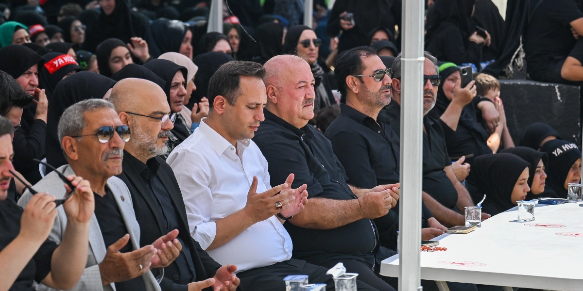Açık havada düzenlenen bir anma töreninde, beyaz bir masa etrafında yan yana oturan ve ellerini dua eder şekilde açmış bir grup erkek görülüyor. Ön planda beyaz gömlekli bir kişi ve yanında siyah kıyafetli katılımcılar yer alıyor. Arka planda ise başörtülü kadınlar ve üzerinde 