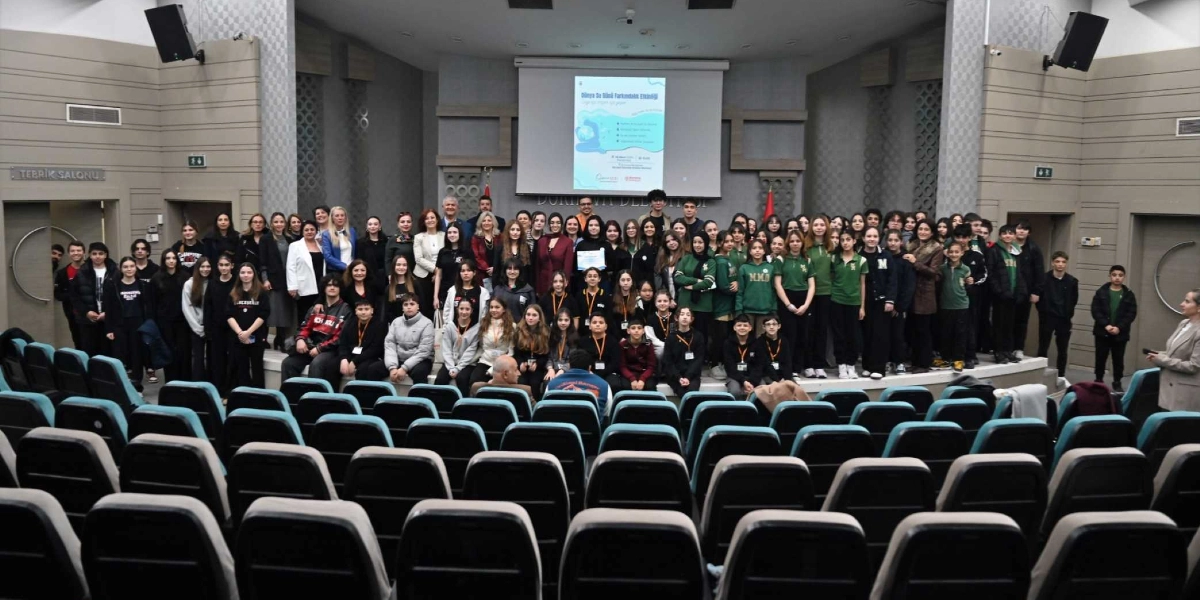 Modern bir konferans salonunda, sahne üzerinde bir araya gelmiş kalabalık bir öğrenci ve yetişkin grubu. Arka planda yer alan projeksiyon perdesinde 