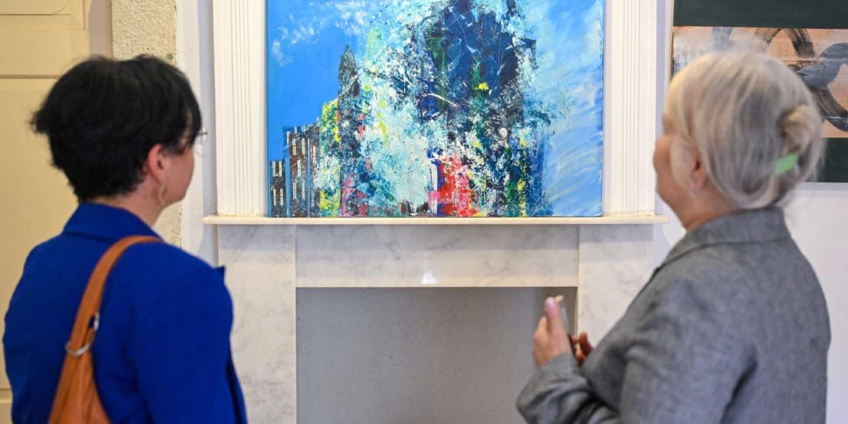 Mavi ceketli ve gri ceketli iki kadın, bir sanat galerisinde mermer şömine rafının üzerinde asılı olan, ağırlıklı olarak mavi tonlarının kullanıldığı soyut bir tabloyu inceliyor.