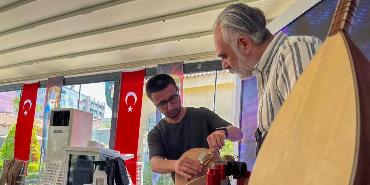 Atölye ortamında, bir bağlama ustası elindeki enstrümanın gövdesine hassas bir işlem uygularken, yanındaki kişi onu dikkatle izliyor. Arka planda camların önünde asılı Türk bayrakları görülüyor.