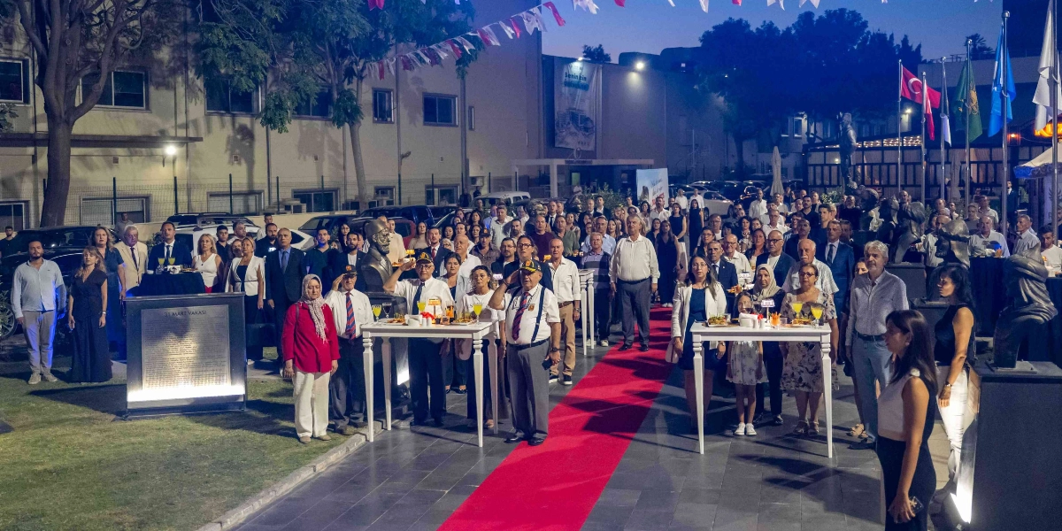Açık havada düzenlenen akşam vakti gerçekleşen resepsiyonda, kırmızı halı üzerinde duran kalabalık bir grup insan saygı duruşunda bulunuyor. Ön planda, üzerinde içecek ve atıştırmalıkların bulunduğu beyaz masaların yanında, askeri üniformalı ve şapkalı gaziler selam duruşu yaparken, arkalarında resmi kıyafetli davetliler yer alıyor. Arka planda Türk bayrakları ve çeşitli flamalar dalgalanırken, alanın sol tarafında üzerinde 