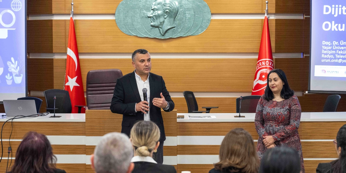 Resmi bir toplantı salonunda, kürsüde konuşma yapan siyah ceketli bir erkek ve yanında duran desenli elbiseli bir kadın katılımcılara hitap ediyor. Arka planda duvarda Atatürk kabartması, iki yanında Türk bayrağı ve Bornova Belediyesi flaması bulunuyor. Yanlardaki projeksiyon ekranlarında dijital okuryazarlık konulu bir sunumun detayları yer alıyor.