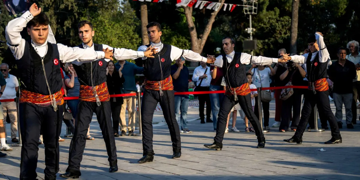 Açık havada düzenlenen bir etkinlikte, geleneksel kıyafetler giymiş beş genç erkek yan yana dizilerek el ele tutuşmuş ve halay çekiyor. Dansçılar beyaz gömlek, üzerinde Türk bayrağı arması bulunan lacivert yelek, koyu renk pantolon ve bellerinde renkli, desenli kuşaklar taşıyor. Arka planda etkinliği izleyen bir kalabalık ve ağaçlar yer alıyor.