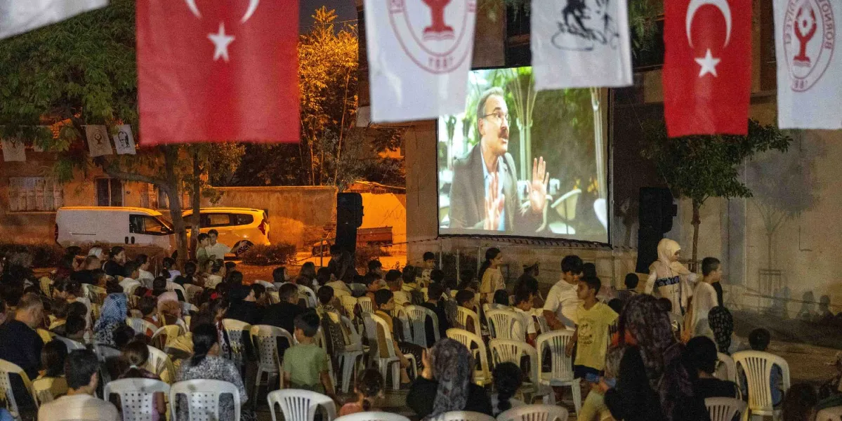 Açık havada kurulan dev bir perdede film izleyen kalabalık bir grup insan. Gece vakti çekilen görüntüde, izleyiciler beyaz plastik sandalyelere oturmuş, perdede konuşan orta yaşlı, gözlüklü bir erkek oyuncuyu izliyor. Alanın üzerinde Türk bayrakları ve Bornova Belediyesi logolu flamalar asılı. Arka planda park edilmiş araçlar ve ağaçlar seçiliyor.
