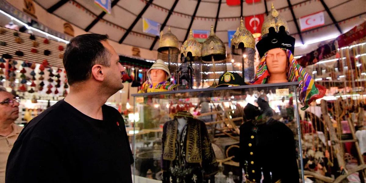 Geleneksel bir çadırın iç mekanında, cam bir vitrinin önünde duran ve sergilenen tarihi miğferleri, yöresel kıyafetleri ve mankenleri inceleyen siyah tişörtlü bir adam. Arka planda Türk bayrağı ve çeşitli Türk devletlerinin bayrakları asılıdır.