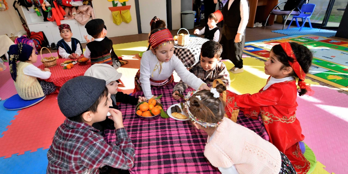 Anaokulu sınıfında geleneksel kıyafetler giymiş çocuklar, üzerinde mandalina ve sarma bulunan yer sofralarının etrafında oturarak Yerli Malı Haftası'nı kutluyor. Bazı çocuklar yöresel başlıklar takarken, bir kız çocuğu elindeki yiyeceği arkadaşına uzatıyor, diğer çocuklar ise masadaki yiyeceklerle ilgileniyor.