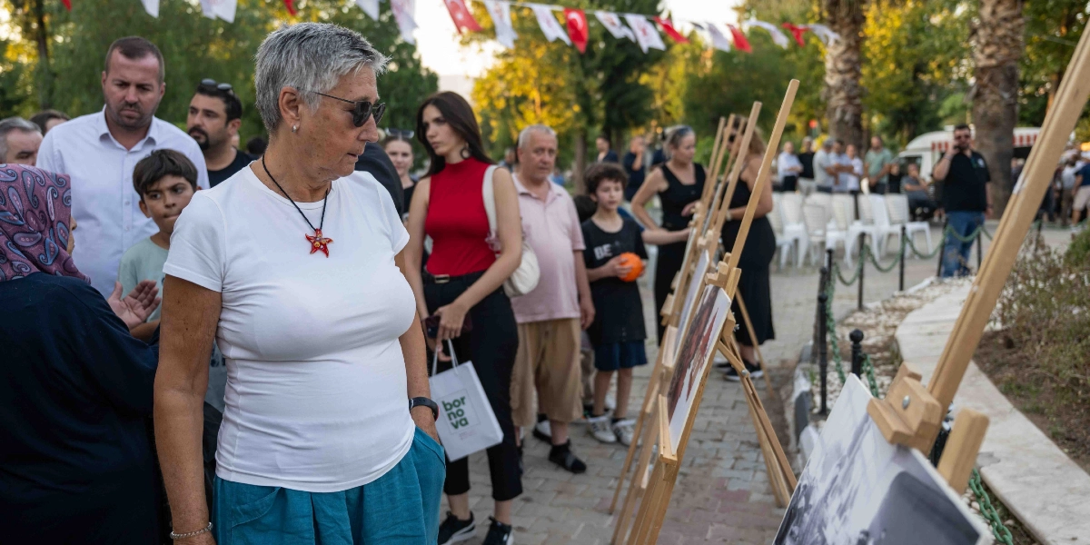 Açık havada düzenlenen bir anma etkinliğinde, insanlar ahşap şövaleler üzerine yerleştirilmiş fotoğrafları inceliyor. Ön planda kısa gri saçlı, beyaz tişörtlü ve güneş gözlüklü bir kadın, yanında başörtülü bir kadın ve arkasında çeşitli yaşlardan katılımcılarla birlikte sergilenen fotoğraflara bakıyor. Arka planda ağaçlar, asılı Türk bayrakları ve oturma düzeni görülürken, katılımcılardan birinin elinde üzerinde 