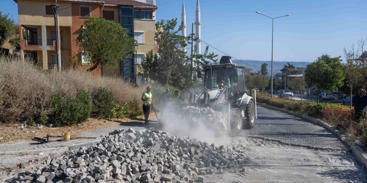 Bornova Belediyesi ekipleri, bir iş makinesi yardımıyla yol kenarındaki parke taşlarını sökerek onarım çalışması yapıyor. Çalışma sırasında toz bulutu yükselirken, görevli personel yol kenarında bekliyor. Arka planda apartmanlar ve bir cami minaresi görülüyor.