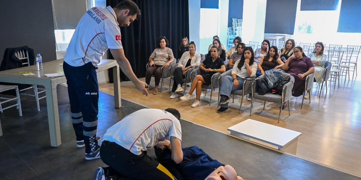 Bornova Belediyesi ambulans üniforması giyen bir sağlık görevlisi, bir grup kadına yerde yatan bir manken üzerinde temel yaşam desteği ve kalp masajı eğitimi veriyor. Sağlık görevlisi, mankenin başında diz çökmüş olan diğer bir görevliyi yönlendirirken, sandalyelerde oturan katılımcılar dikkatle uygulamayı izliyor.