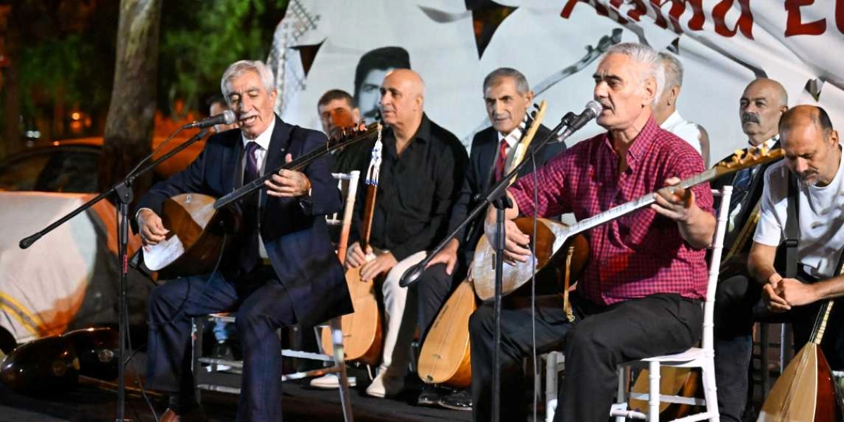 Açık havada düzenlenen bir etkinlikte, ellerinde bağlamalarıyla oturan ve mikrofon başında performans sergileyen halk ozanları. Ön planda, biri koyu renk takım elbiseli, diğeri ise kırmızı kareli gömlekli iki ozan bağlama çalarken, arkalarında onları dinleyen veya eşlik eden diğer müzisyenler yer alıyor. Arka planda ise üzerinde kısmen görünen yazılar ve insan silüetleri bulunan beyaz bir fon bulunuyor.