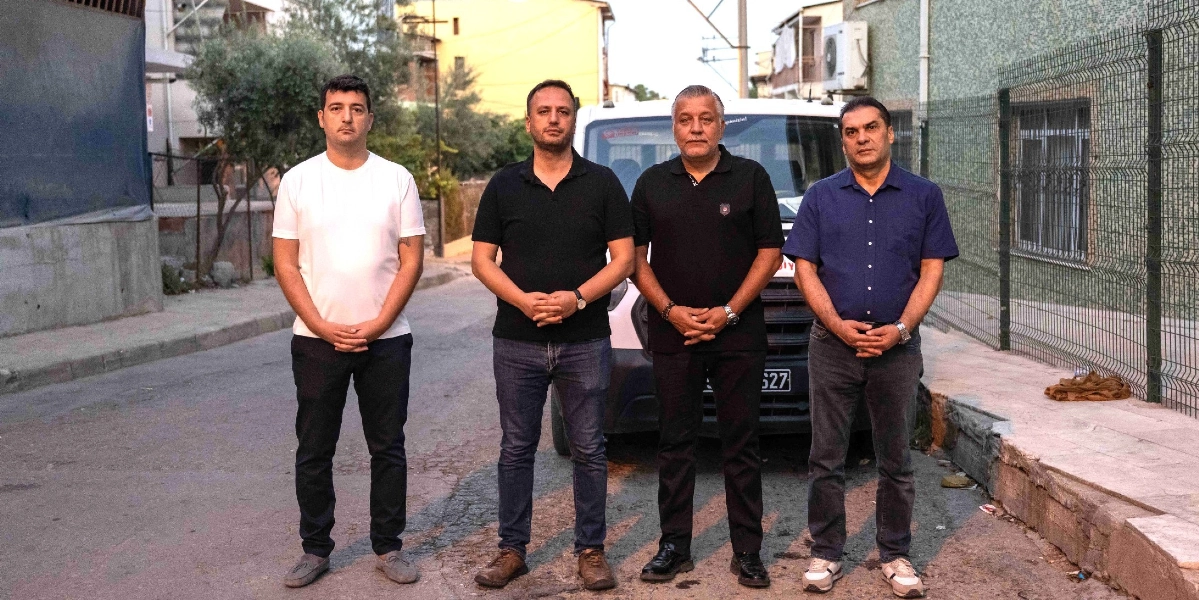 Dört erkek, sabahın erken saatlerinde bir sokakta yan yana durarak kameraya bakıyor. En soldaki kişi beyaz tişört ve koyu renk pantolon giyerken, yanındaki iki kişi siyah polo yaka tişört ve kot pantolon, en sağdaki kişi ise lacivert kısa kollu gömlek ve gri pantolon giyiyor. Arkalarında beyaz bir hizmet aracı, sağ tarafta ise tel örgülü bir bahçe duvarı bulunuyor.