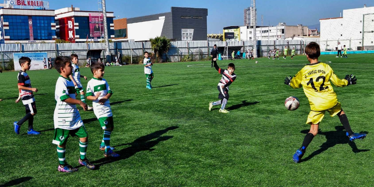 Açık havada, suni çim kaplı bir futbol sahasında düzenlenen çocuk futbol turnuvasından bir an. Sarı formasıyla kaleci, kendisine doğru gelen topu uzaklaştırmaya çalışırken, beyaz ve yeşil çizgili formalar giyen diğer çocuklar oyun alanında pozisyon alıyor. Arka planda binalar ve saha çevresini saran metal çitler görülüyor.