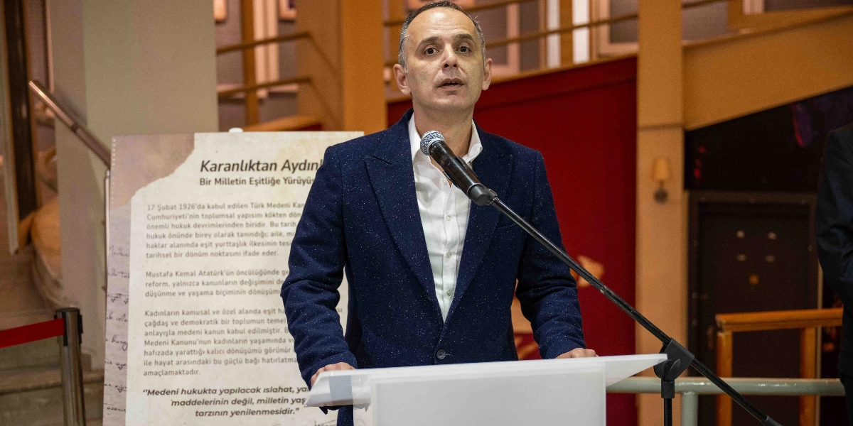 Koyu renkli bir ceket ve beyaz gömlek giymiş bir erkek, kürsüde mikrofon başında konuşma yapıyor. Arka planda üzerinde 