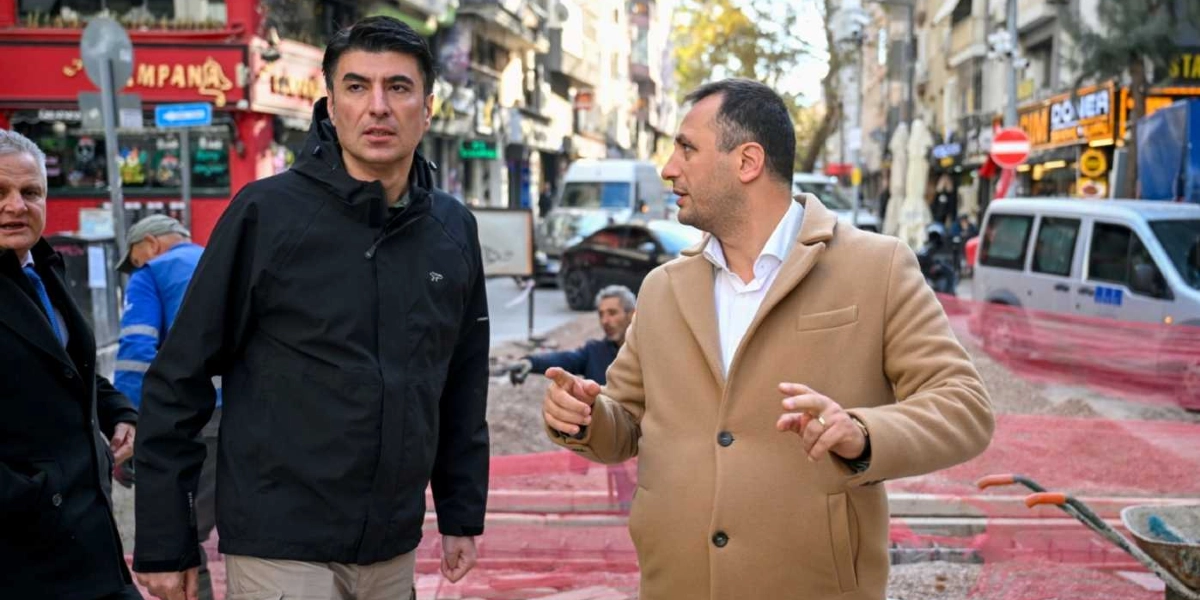 Bornova'da bir cadde üzerinde devam eden altyapı çalışmalarını inceleyen iki yetkili, çevreye bakarak konuşuyor. Siyah ceketli bir kişi ve bej rengi kabanlı bir kişi, arka planda kazı alanı ve iş makinelerinin bulunduğu çalışma sahasında incelemelerde bulunuyor.