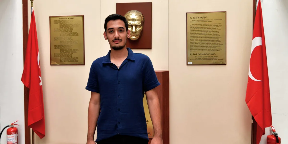 Kısa saçlı, hafif sakallı ve lacivert kısa kollu bir gömlek giyen genç bir erkek, arka planda Atatürk rölyefi, sağında ve solunda Türk bayrakları, yan duvarlarda ise İstiklal Marşı ve Gençliğe Hitabe metinlerinin bulunduğu bir odada duruyor.