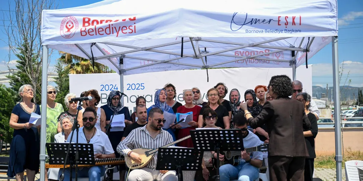 Bornova Belediyesi tarafından düzenlenen bir etkinlikte, beyaz bir çadırın altında şarkı söyleyen bir koro ve onlara eşlik eden müzisyenler yer alıyor. Ön planda bağlama ve gitar çalan müzisyenler, arkalarında ise ellerinde notalarla şarkı söyleyen, çoğunluğu kadınlardan oluşan koro grubu bulunuyor. Grubun önünde, sırtı dönük bir şekilde koroyu yöneten bir şef yer alıyor. Çadırın üzerinde 
