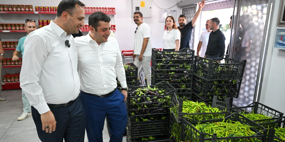 Beyaz gömlekli iki erkek, bir marketin içinde yan yana durarak gülümseyerek sohbet ediyor. Arka planda raflarda kavanozlu ürünler ve taze sebzelerle dolu siyah plastik kasalar bulunuyor. Çevredeki diğer insanlar da marketin içinde duruyor, bir kişi elini havaya kaldırarak selam veriyor.